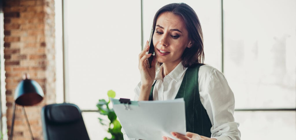 Businesswoman in formalwear multitasking in a modern office setting, talking on the phone and reviewing documents with professionalism.