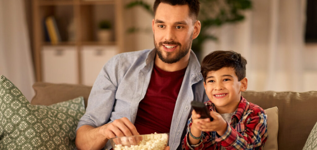 family, childhood, fatherhood, technology and people concept - happy father and little son with popcorn watching tv at home in evening
